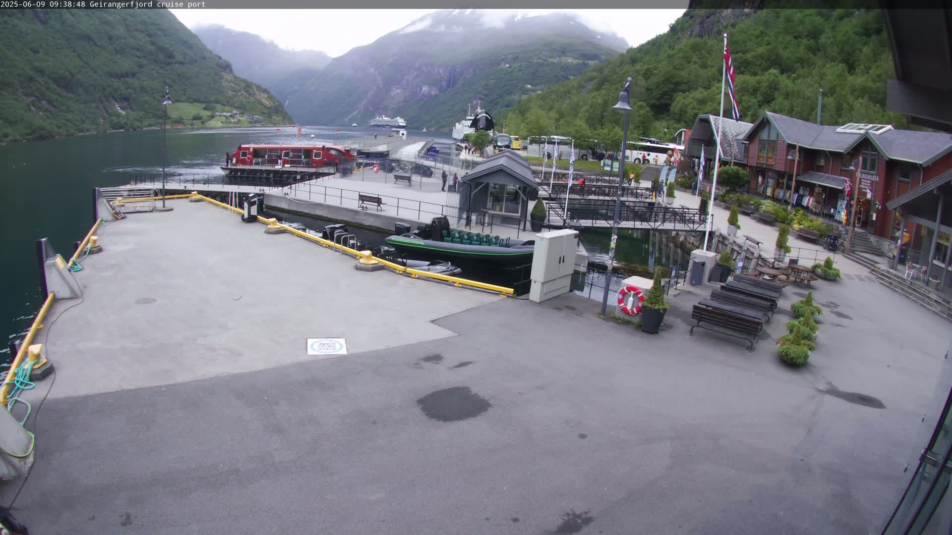 A cruise ship port in a fjord, with overcast weather, shows several small boats docked, a larger red passenger ferry, and buildings with shops.