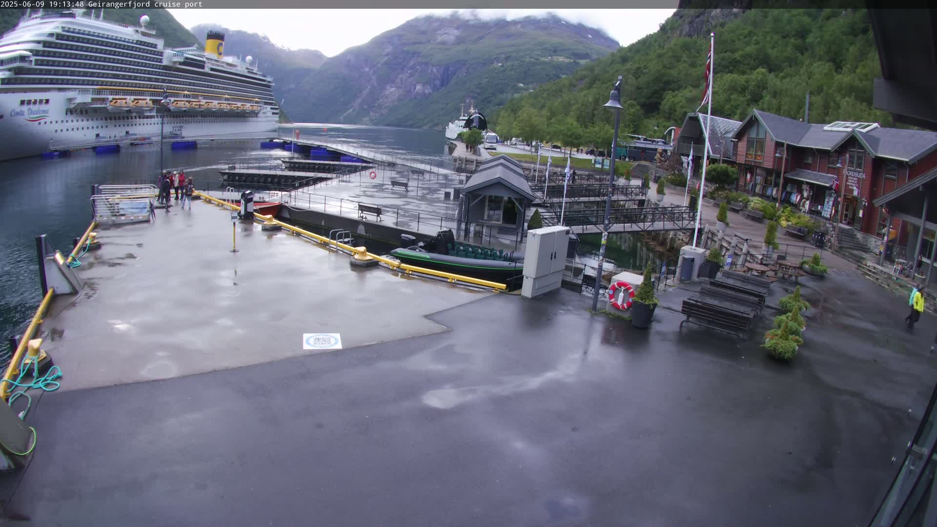 A large cruise ship is docked at a port in a fjord surrounded by mountains, under a rainy sky.