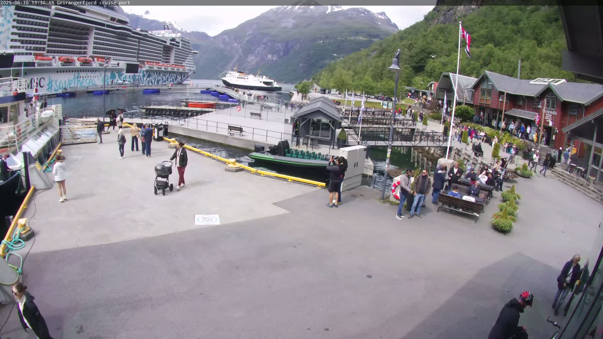 A cruise ship is docked at a port, with many people walking around the area near several buildings and a small boat.