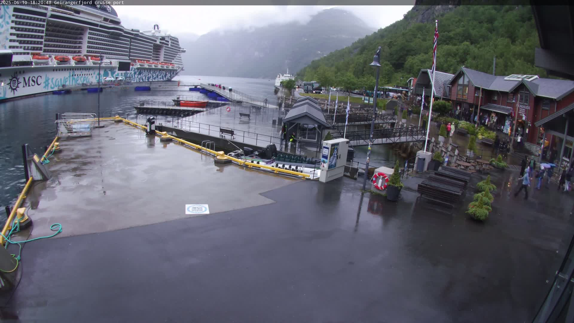 A large cruise ship is docked at a wet, rainy cruise port, with several people walking around the shops and buildings nearby.