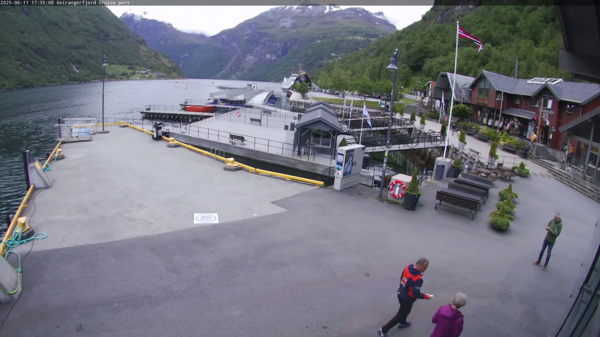 A cruise ship port situated in a fjord, under partly cloudy skies, features a paved area with several buildings, benches, and people walking around.
