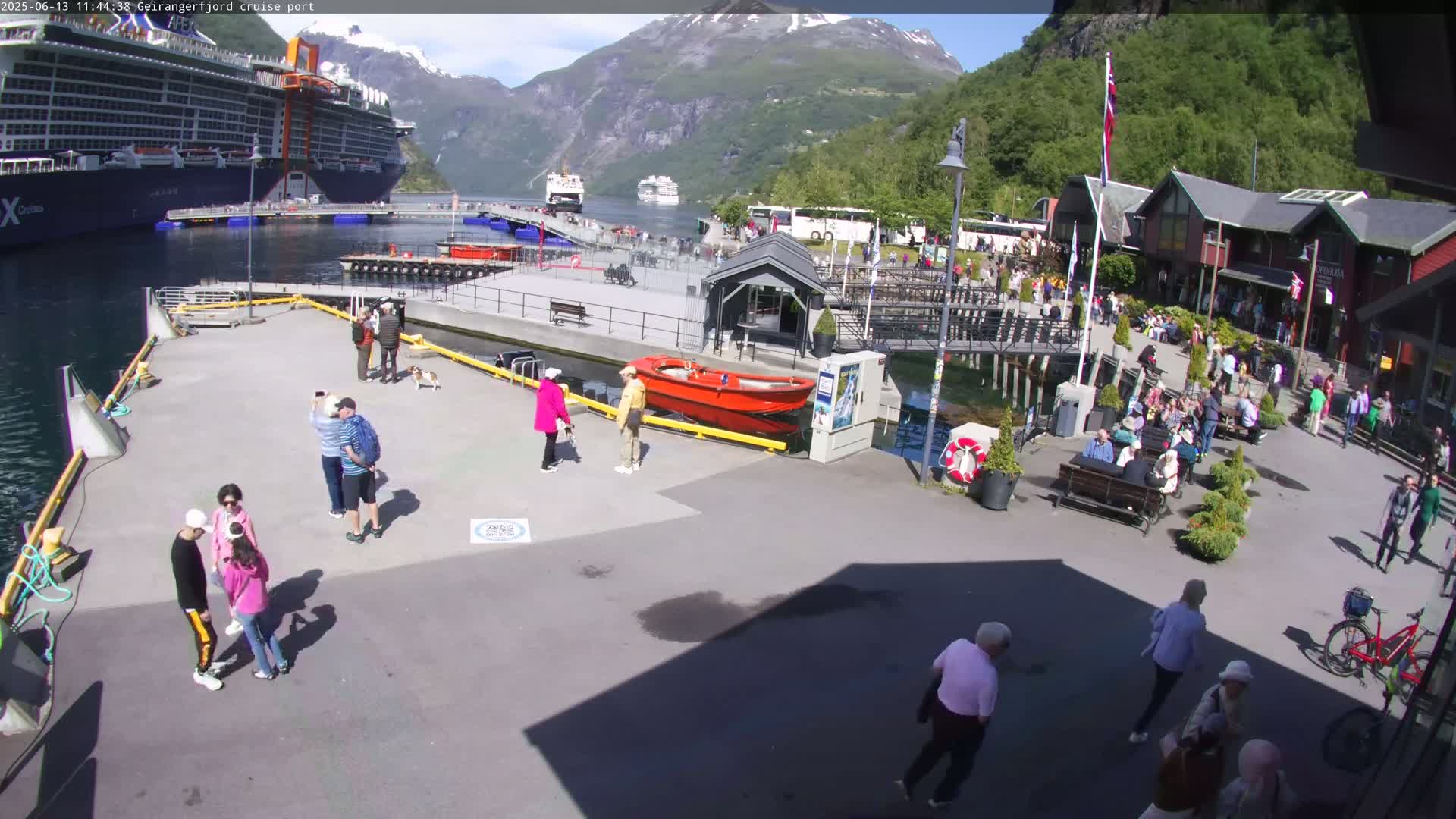 A cruise ship is docked at a port in a fjord, with numerous people milling around the pier on a sunny day.