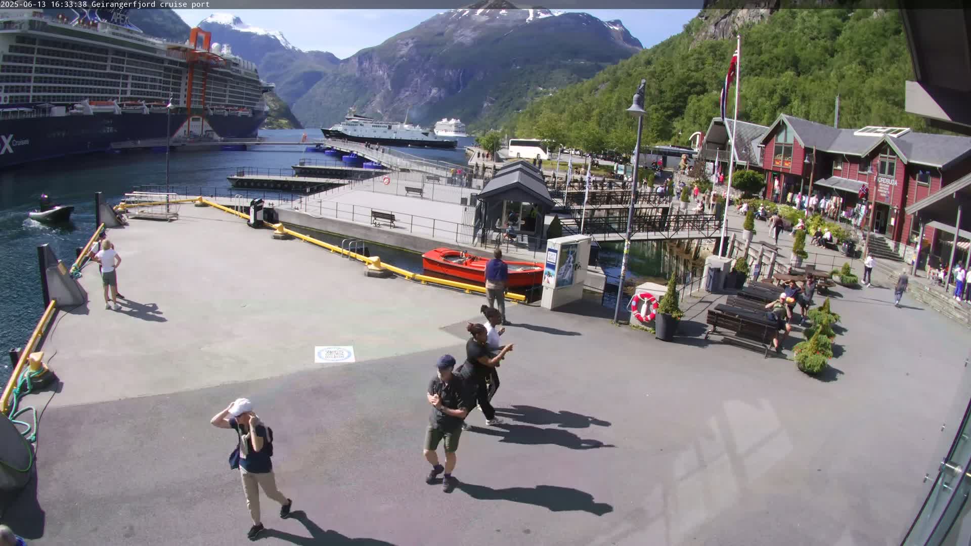 A sunny day at a cruise port shows several people walking around near docked cruise ships and buildings, with mountains in the background.