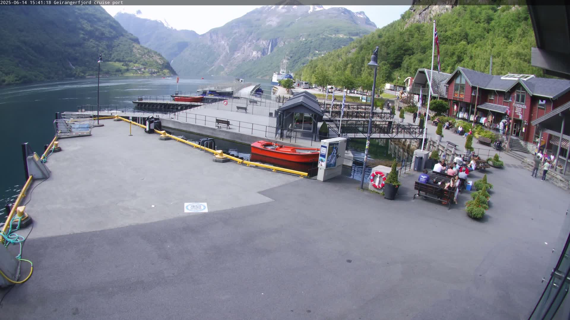 A cruise ship port in a fjord, under sunny skies, features a red motorboat at a dock, several people sitting at outdoor tables, and buildings along the shore with mountains in the background.