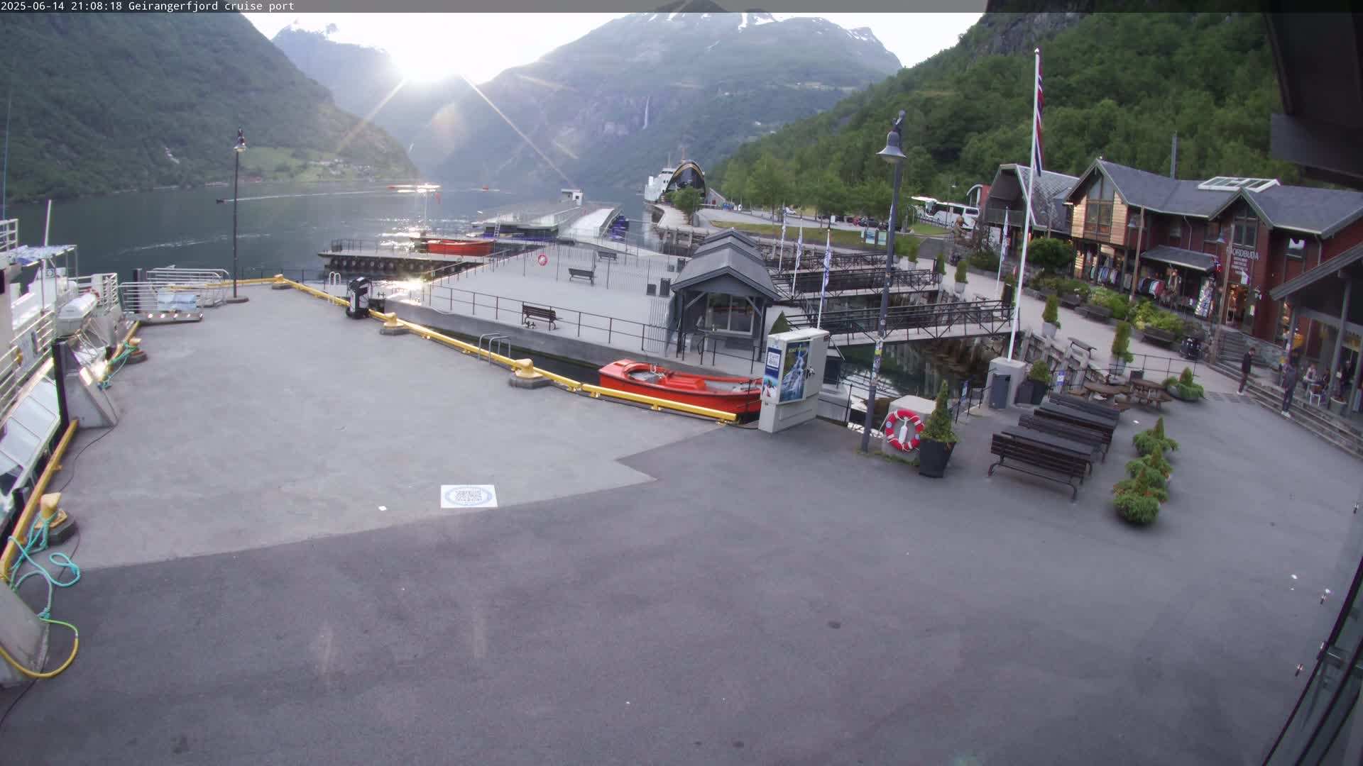 A cruise ship port in a fjord, under sunny skies, features a red motorboat at a dock, several people sitting at outdoor tables, and buildings along the shore with mountains in the background.