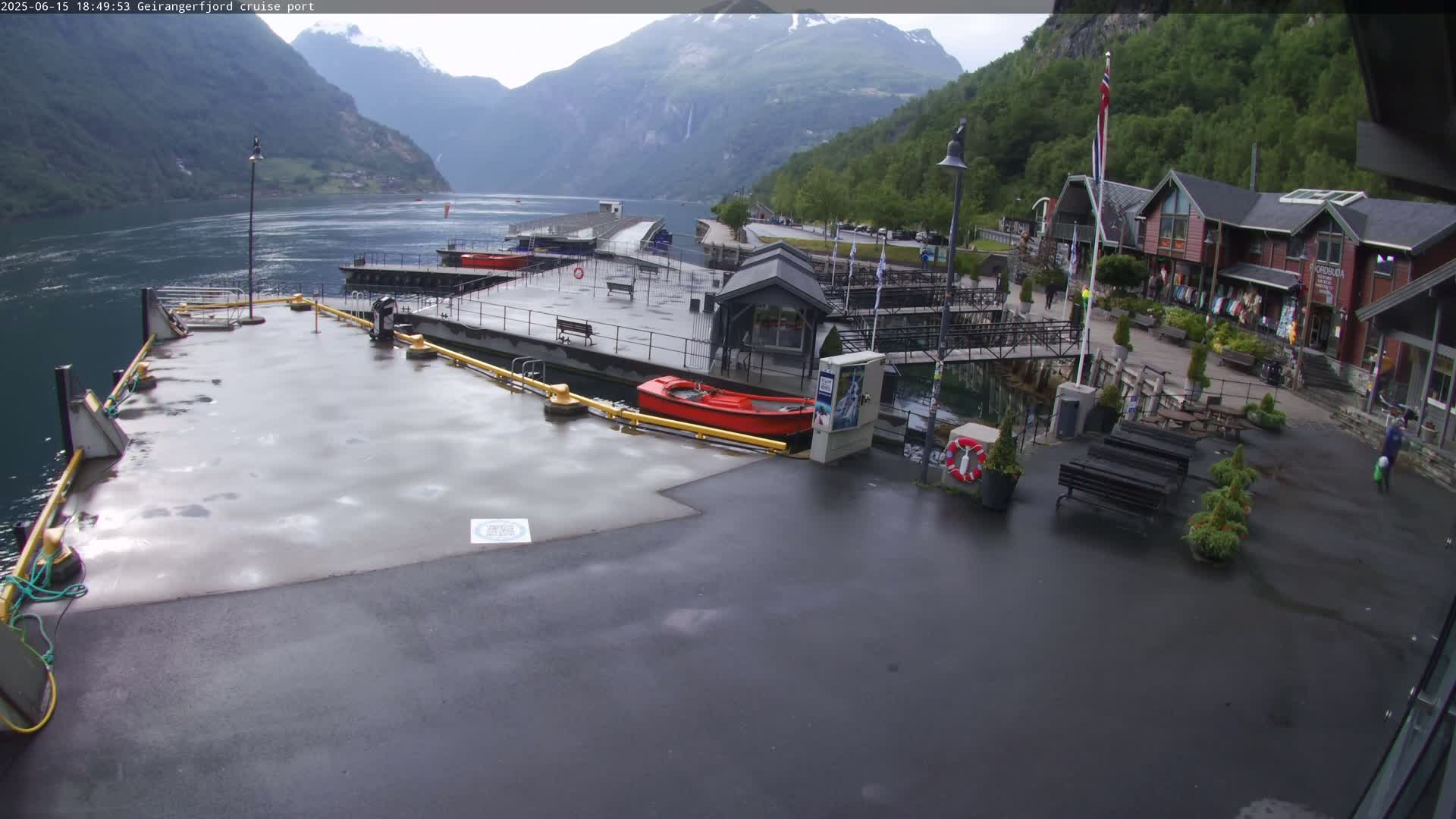A wet cruise ship port with several small buildings, a red rowboat, and a calm body of water surrounded by mountains under an overcast sky.