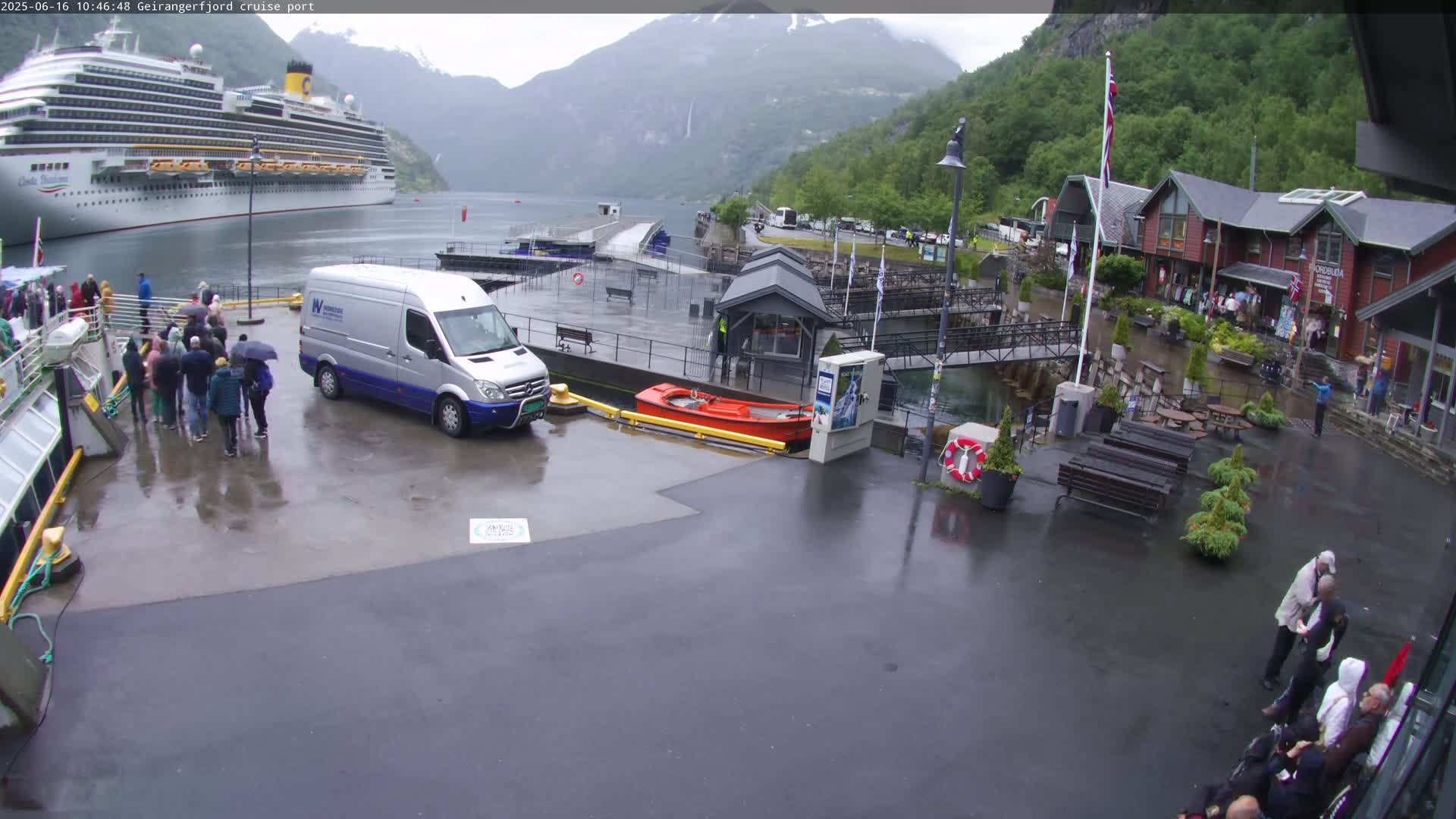 A large cruise ship is docked at a port in a mountainous area during rainy weather; people are disembarking the ship and walking on a wet dock.