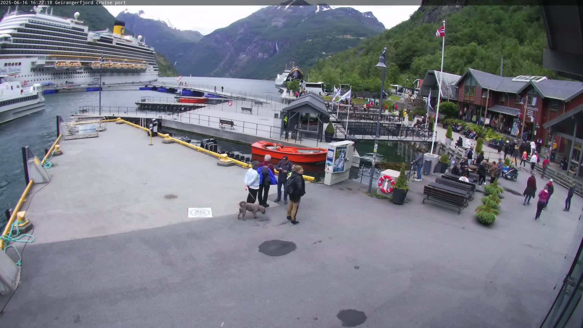 A cruise ship is docked at a port in a fjord surrounded by mountains on an overcast day, with several people walking around the area.