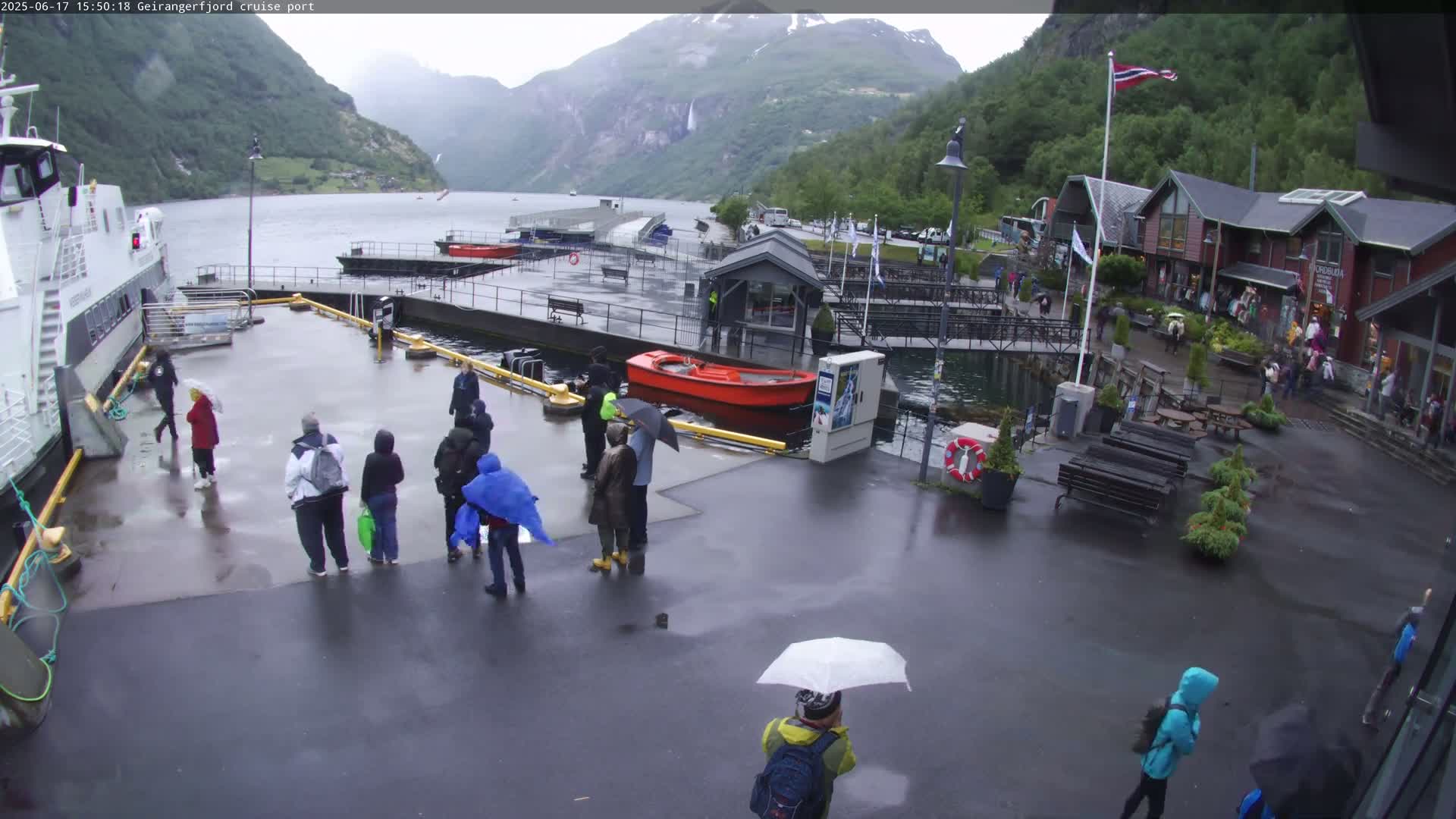 A rainy day at a cruise port shows several people walking around near a docked ferry and various small buildings, with mountains in the background.
