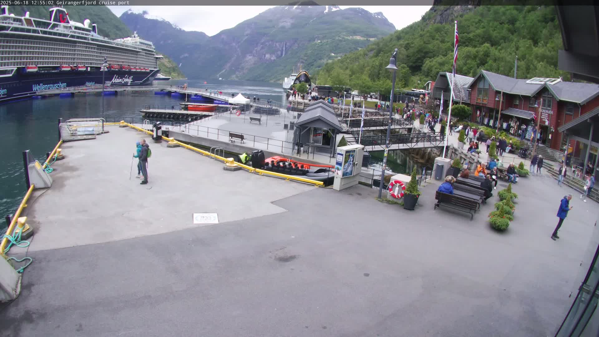 A cruise ship is docked at a port in a fjord surrounded by mountains under a partly cloudy sky, with people walking around the port area and sitting on benches.