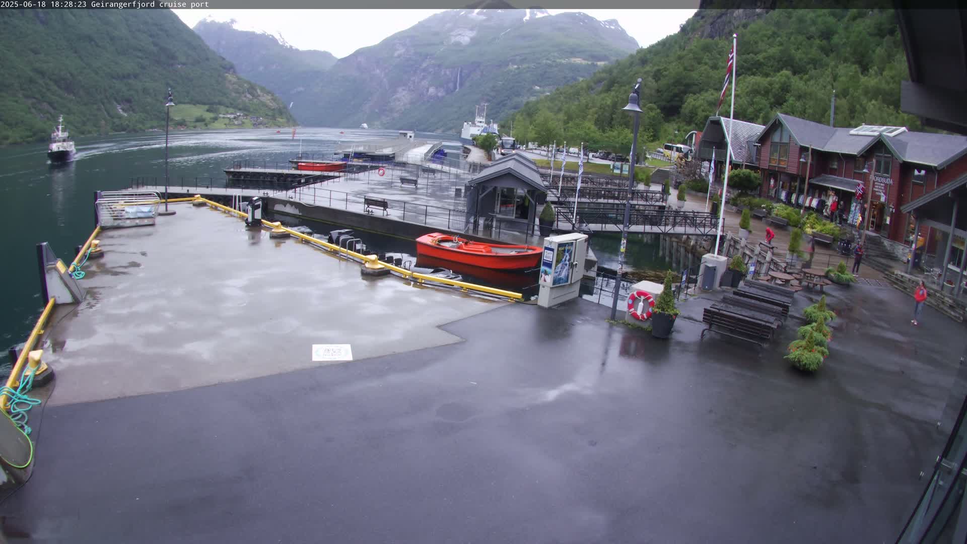 A wet dockside area with a red motorboat, several buildings, and a fjord with mountains in the background under overcast skies.