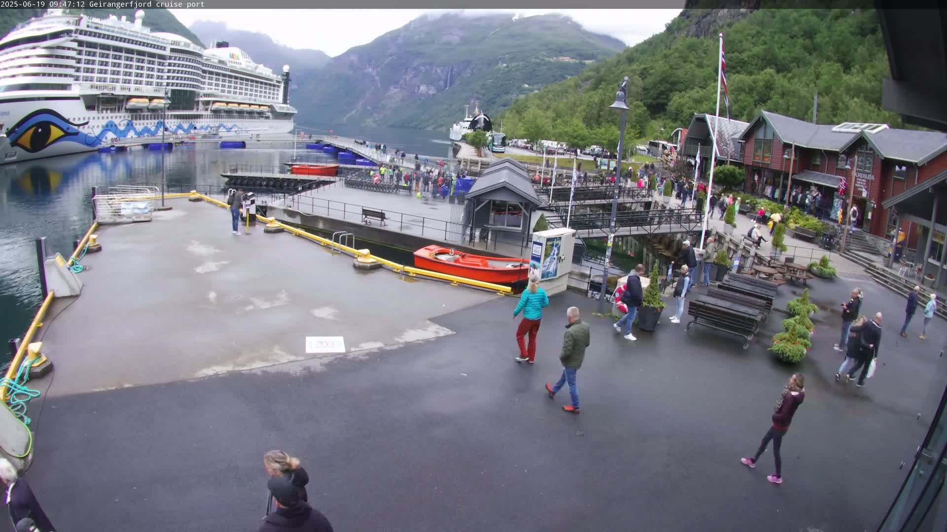 A large cruise ship is docked at a port in a fjord surrounded by green mountains under an overcast sky, with many people walking around the port area.