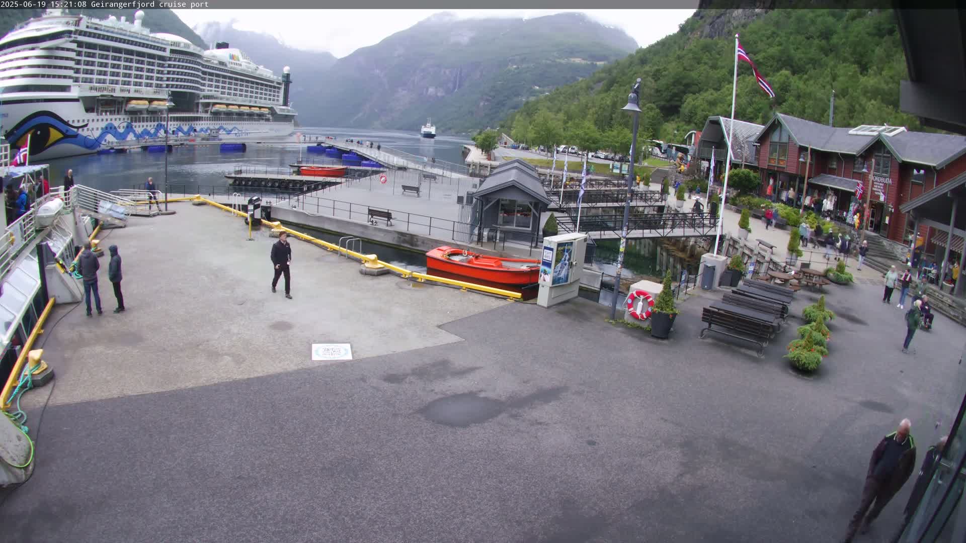A cruise ship is docked at a port in a fjord, with overcast weather and several people walking around the area near several buildings.
