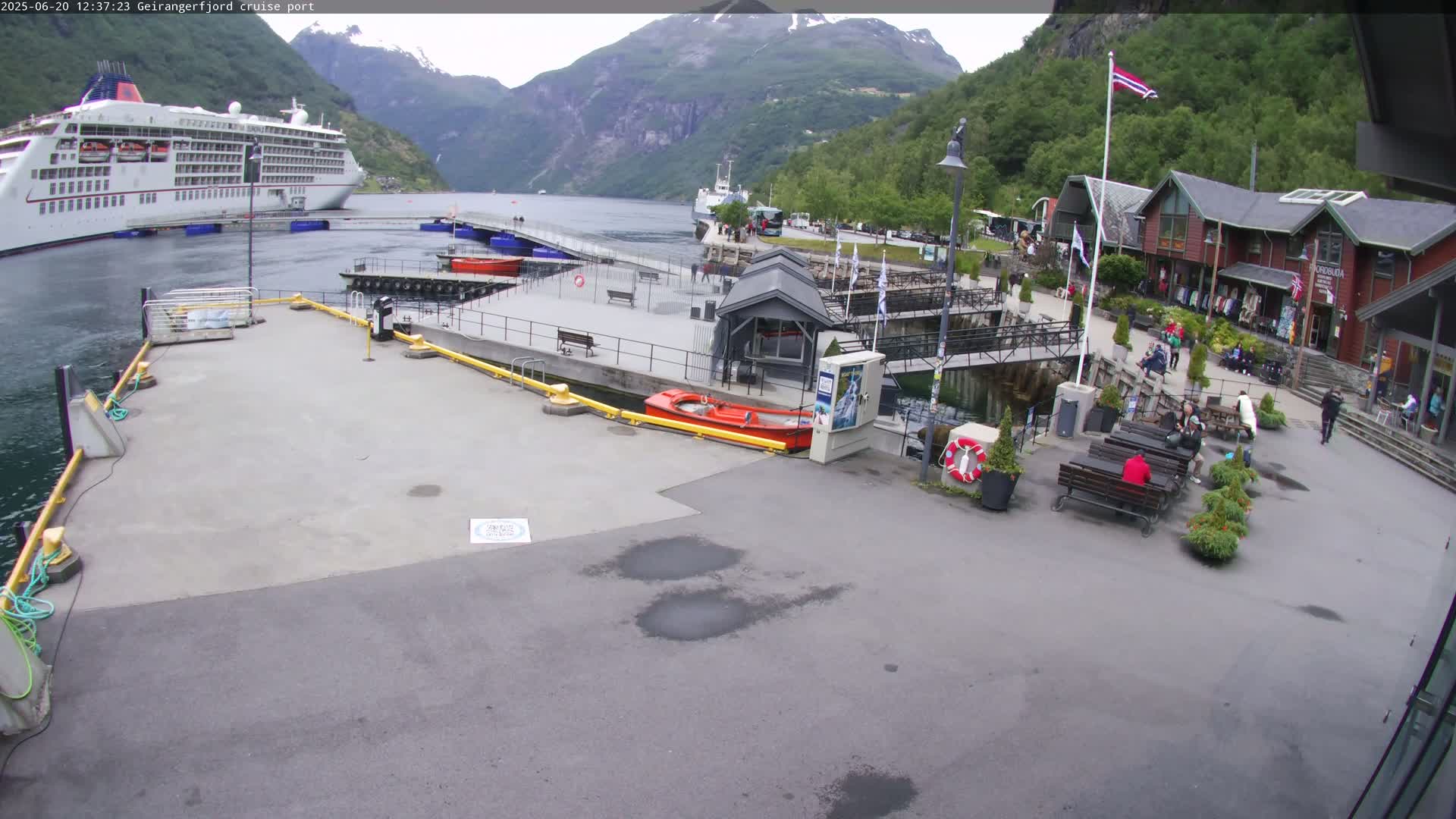 A cruise ship is docked at a port in a fjord, surrounded by mountains and several small buildings with people sitting at outdoor seating areas on a partly cloudy day.