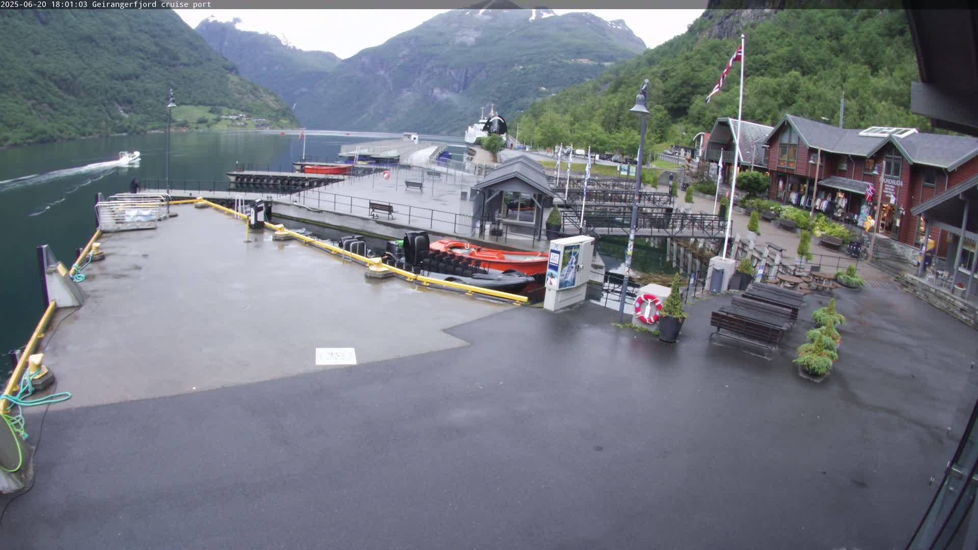 A wet cruise ship port with boats docked, a few buildings, and benches sits nestled in a fjord surrounded by steep, green mountains under an overcast sky.