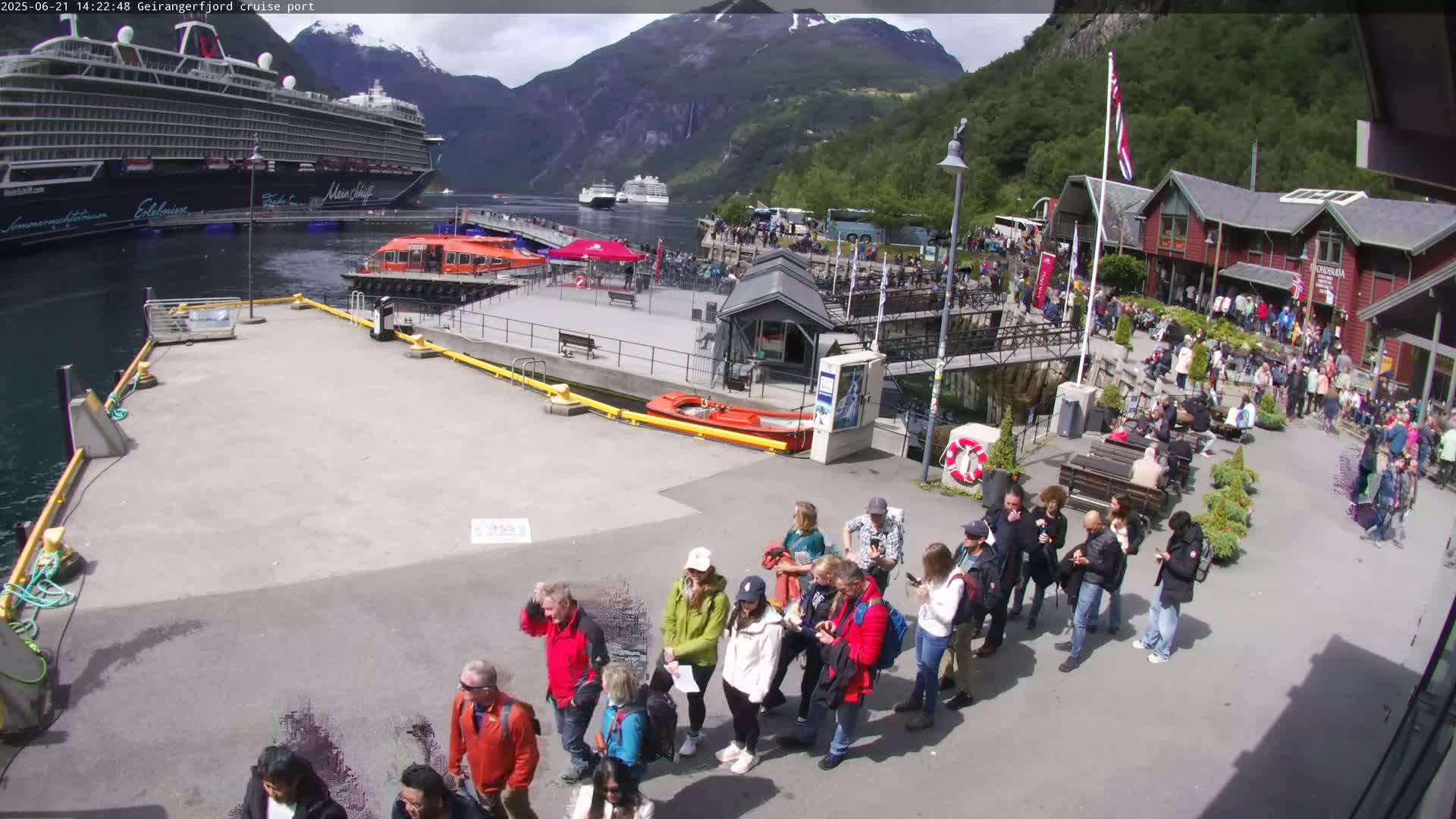 A large cruise ship is docked at a port in a fjord, with many people gathered on the pier and surrounding buildings under partly cloudy skies.
