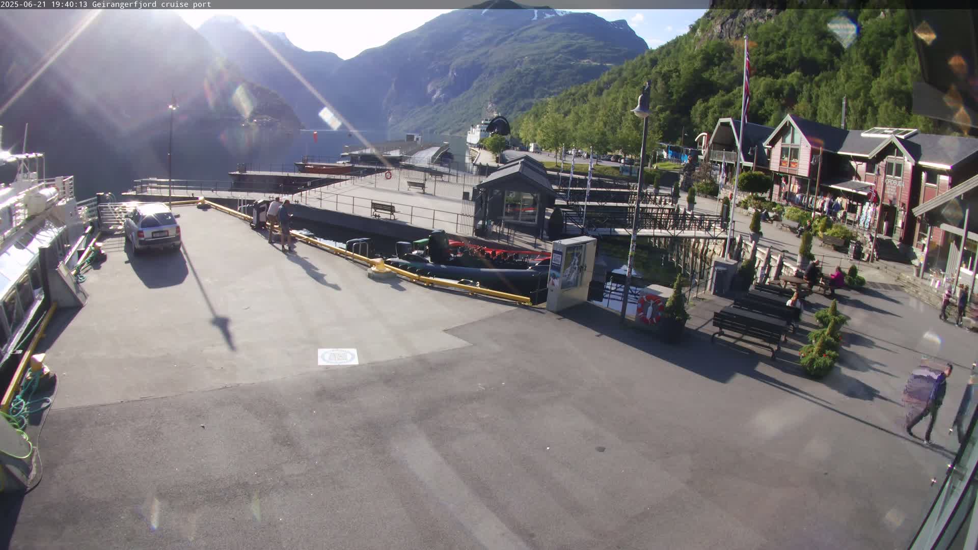 A sunny day at a cruise port nestled between mountains, featuring a car, several people, buildings, boats, and a paved area.