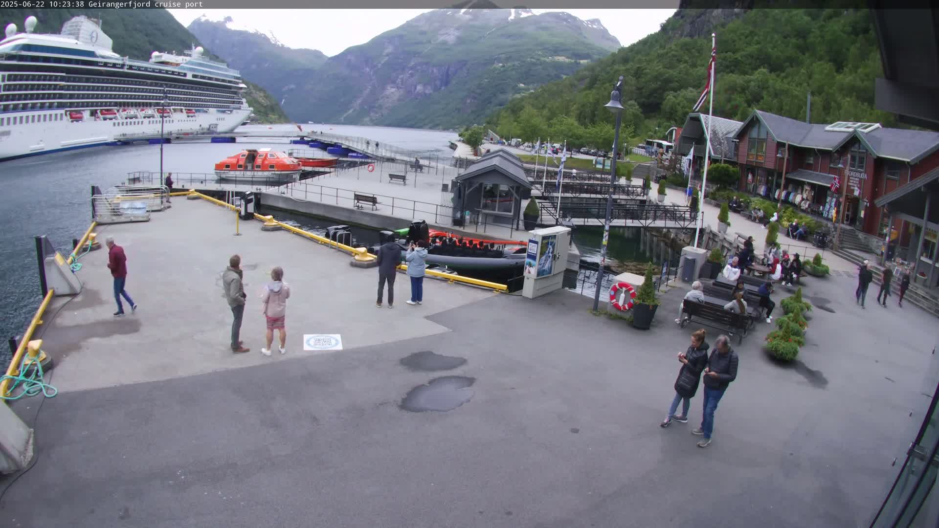 A cruise ship is docked at a port in a fjord surrounded by mountains, with several people milling about the area on a partly cloudy day.
