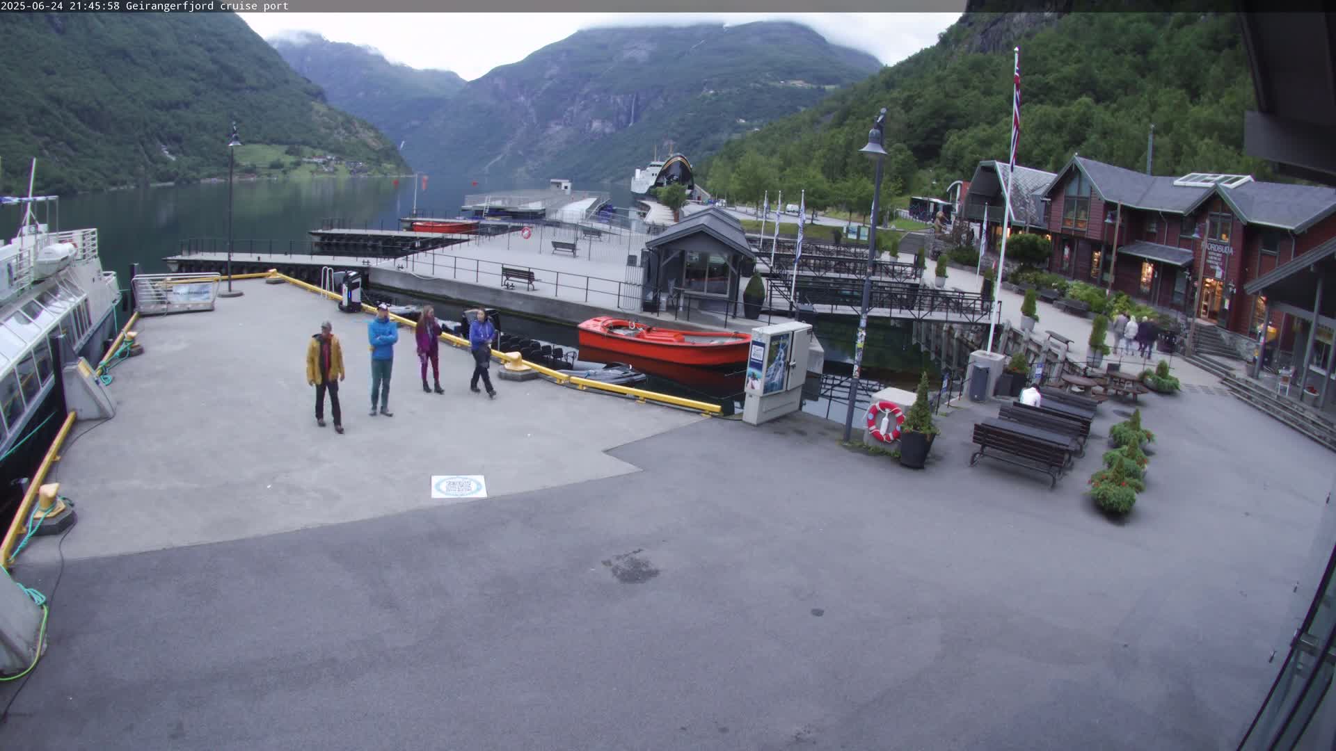 A cruise ship port with several people standing on the dock, a red motorboat, and a calm body of water surrounded by mountains under an overcast sky.