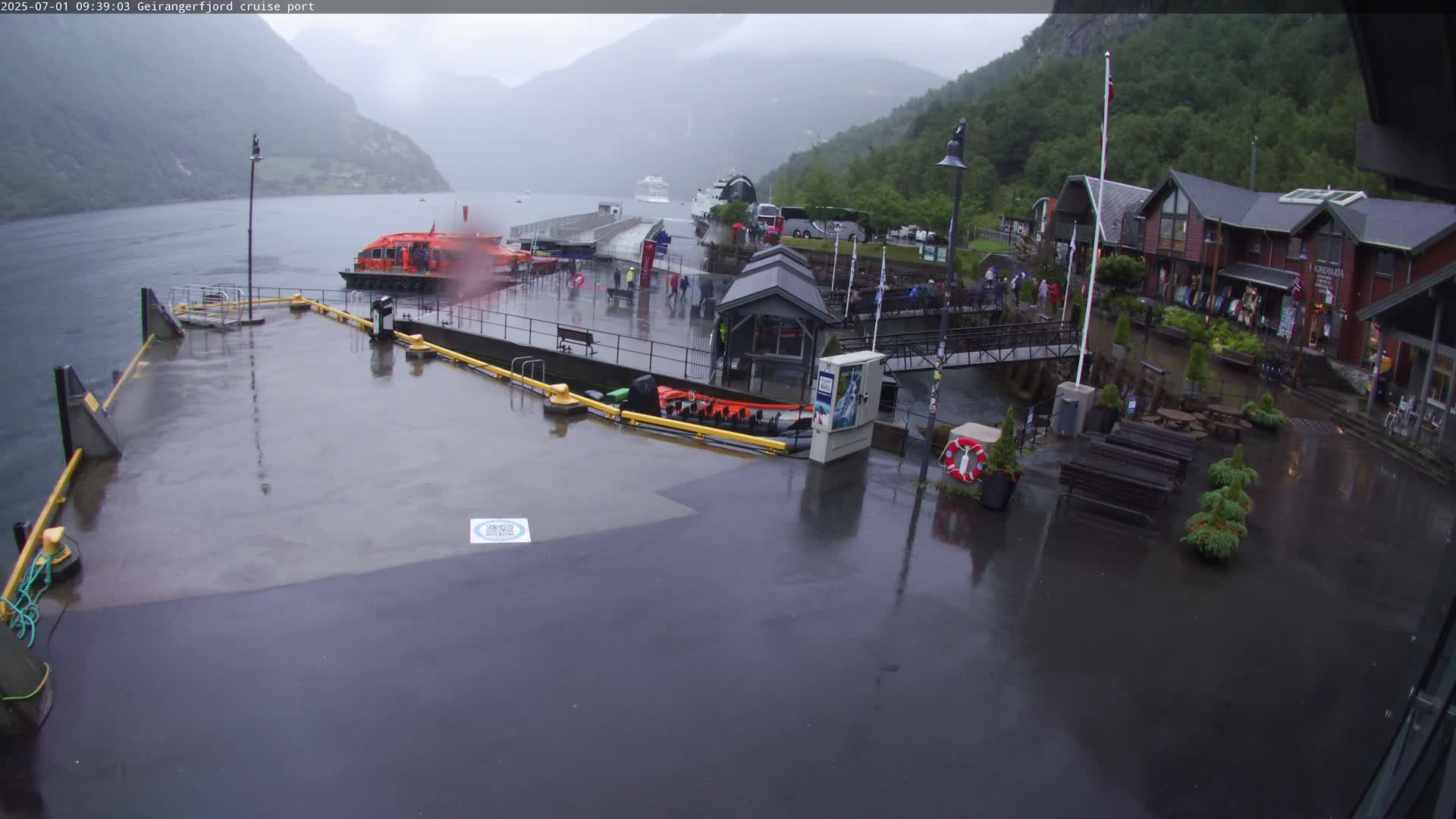 A rain-soaked cruise port features a small orange boat docked near a wet concrete area with several people and buildings visible in the background, nestled between mountains under an overcast sky.