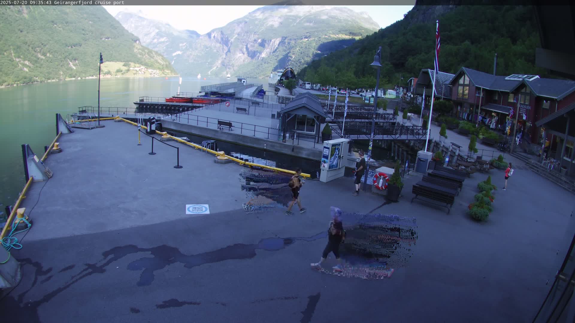 A cruise port sits on a calm body of water next to steep, green mountains under a mostly clear sky.