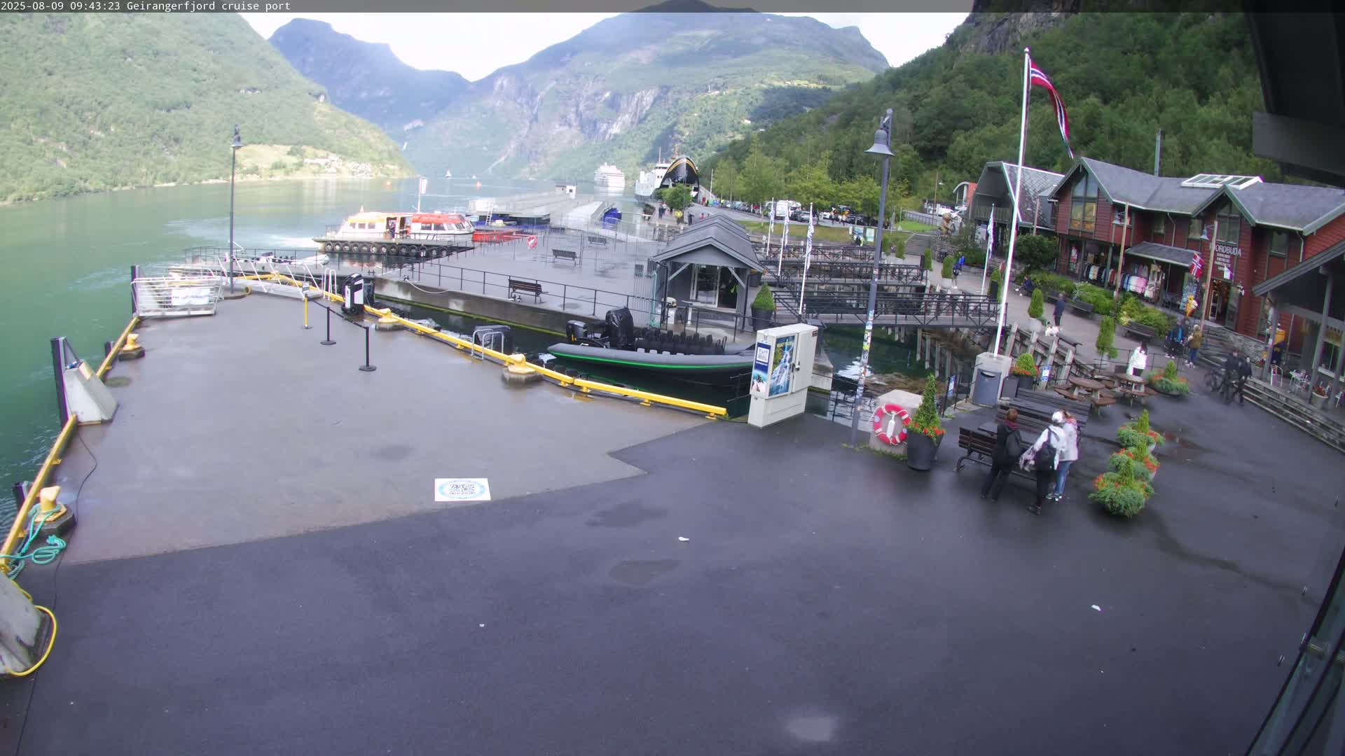 A wet cruise ship port with several small boats and a few people walking around, nestled between steep, green mountains on a cloudy day.