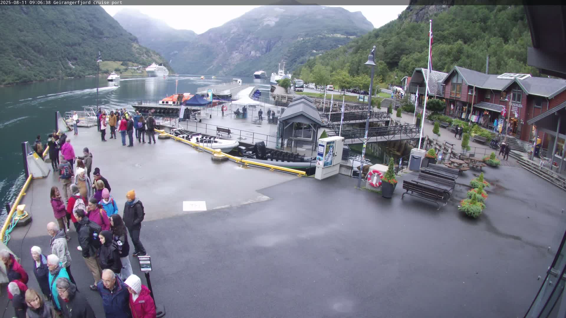A cruise port in a fjord, under an overcast sky, shows numerous people gathered on a dock with boats and shops nearby, and cruise ships visible in the water.