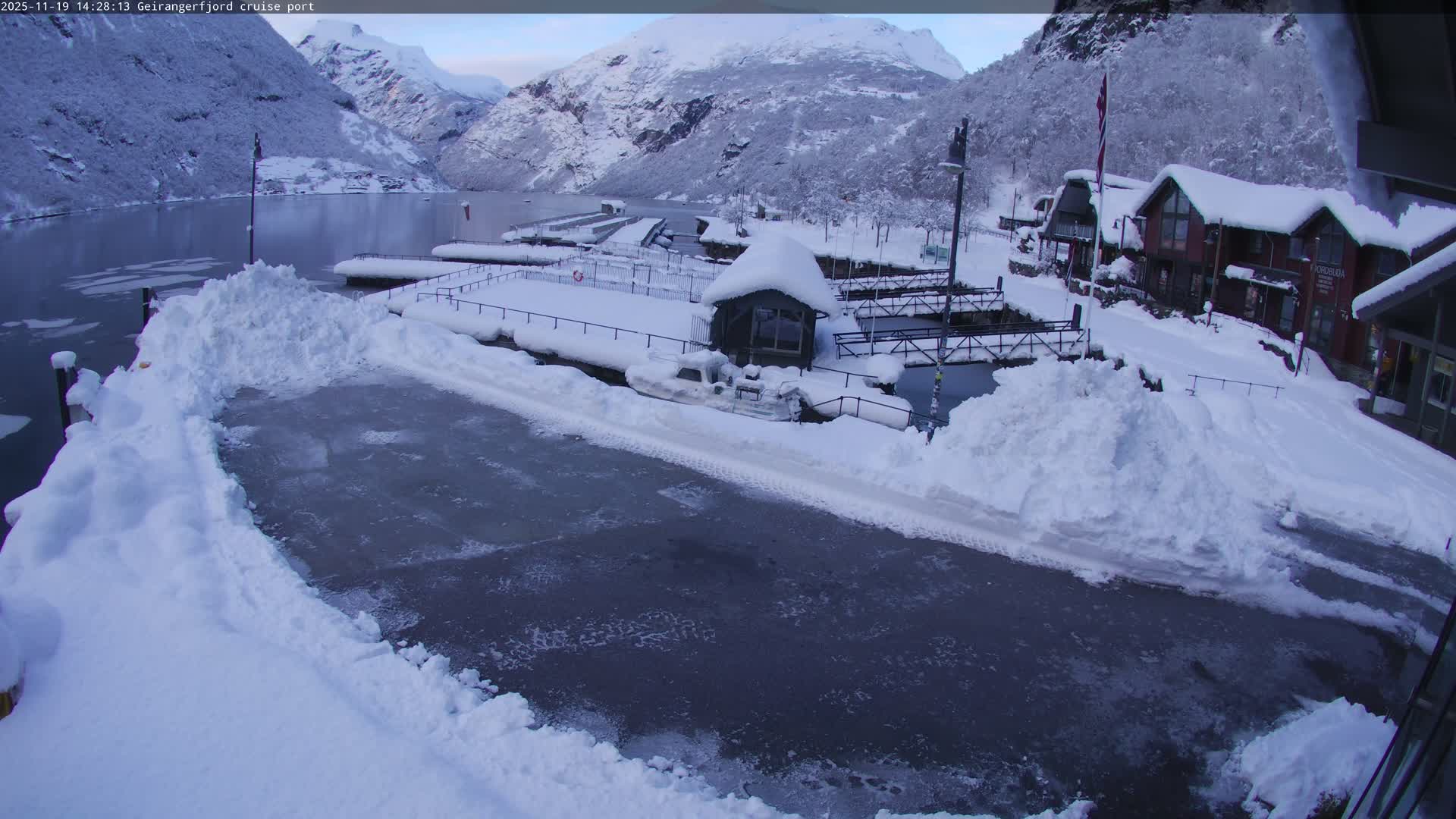 A cruise port in a fjord, under an overcast sky, shows numerous people gathered on a dock with boats and shops nearby, and cruise ships visible in the water.