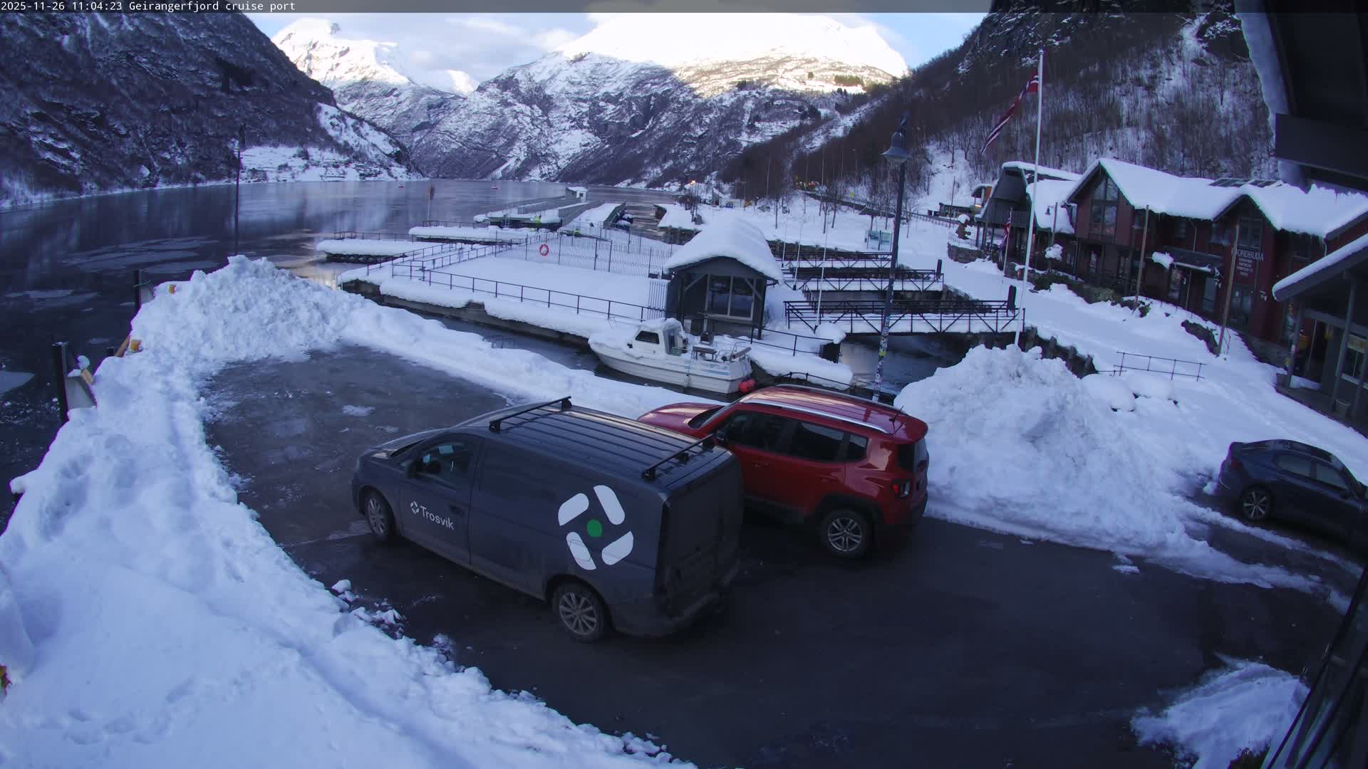 A snow-covered Norwegian fjord village, nestled between towering white mountains, features a partially iced harbor with boats and several winter-clad buildings, all under crisp, partly sunny winter weather.