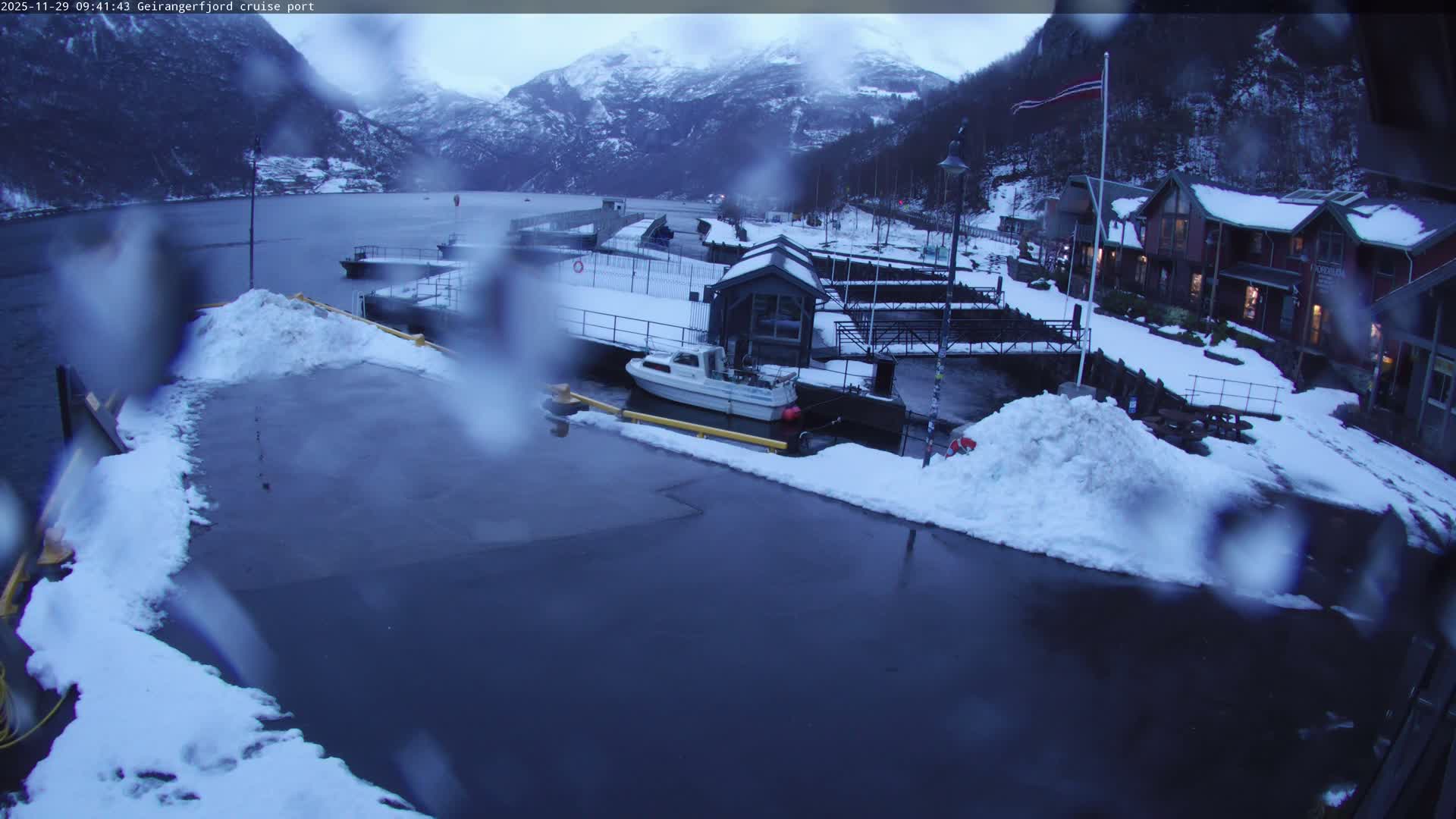 A wintry, overcast day at a fjord port reveals dark water, snow-covered ground and structures including a docked boat, warmly lit buildings, and imposing snow-capped mountains under a grey sky, with the view slightly obscured by water droplets on the lens.