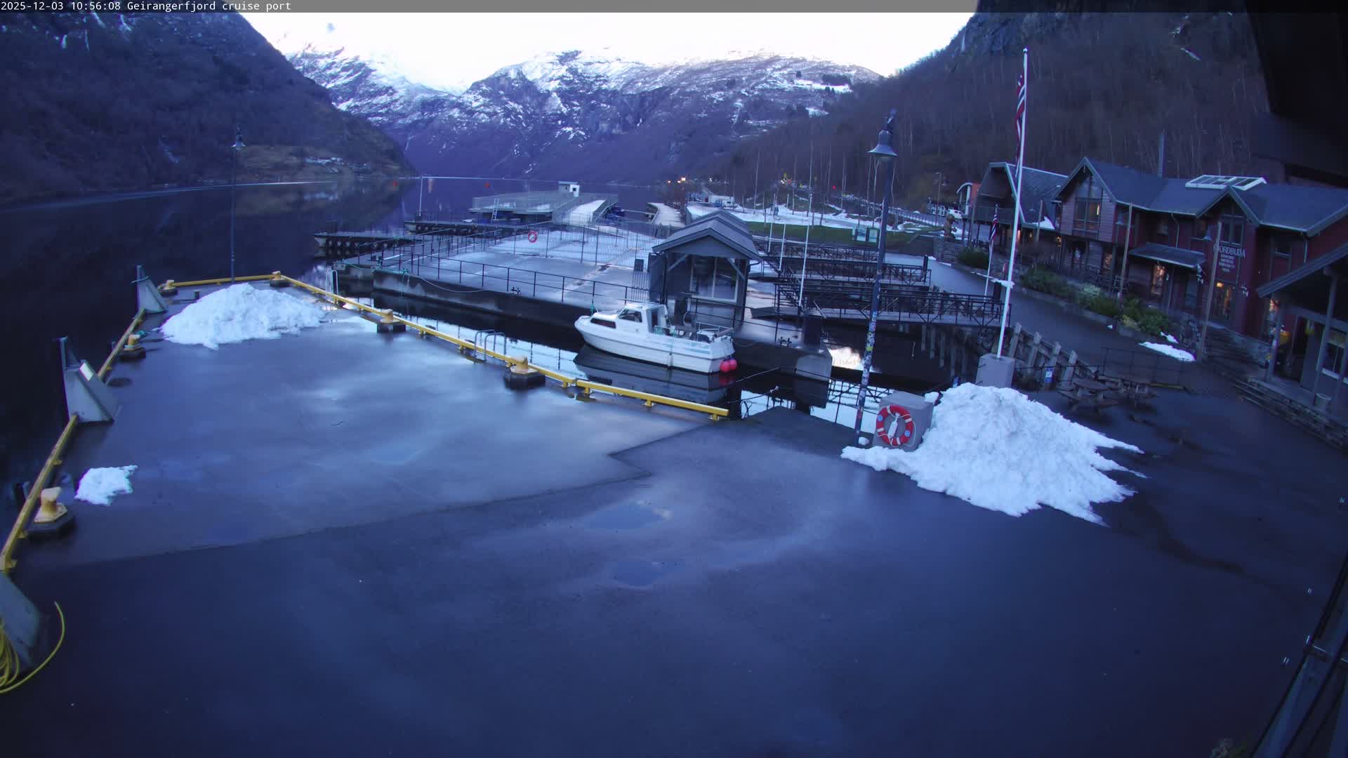 A serene winter landscape depicts a fjord port with dark, calm water reflecting snow-capped mountains, featuring a small boat docked at a pier adorned with piles of snow, and several waterside buildings under an overcast and cold sky.