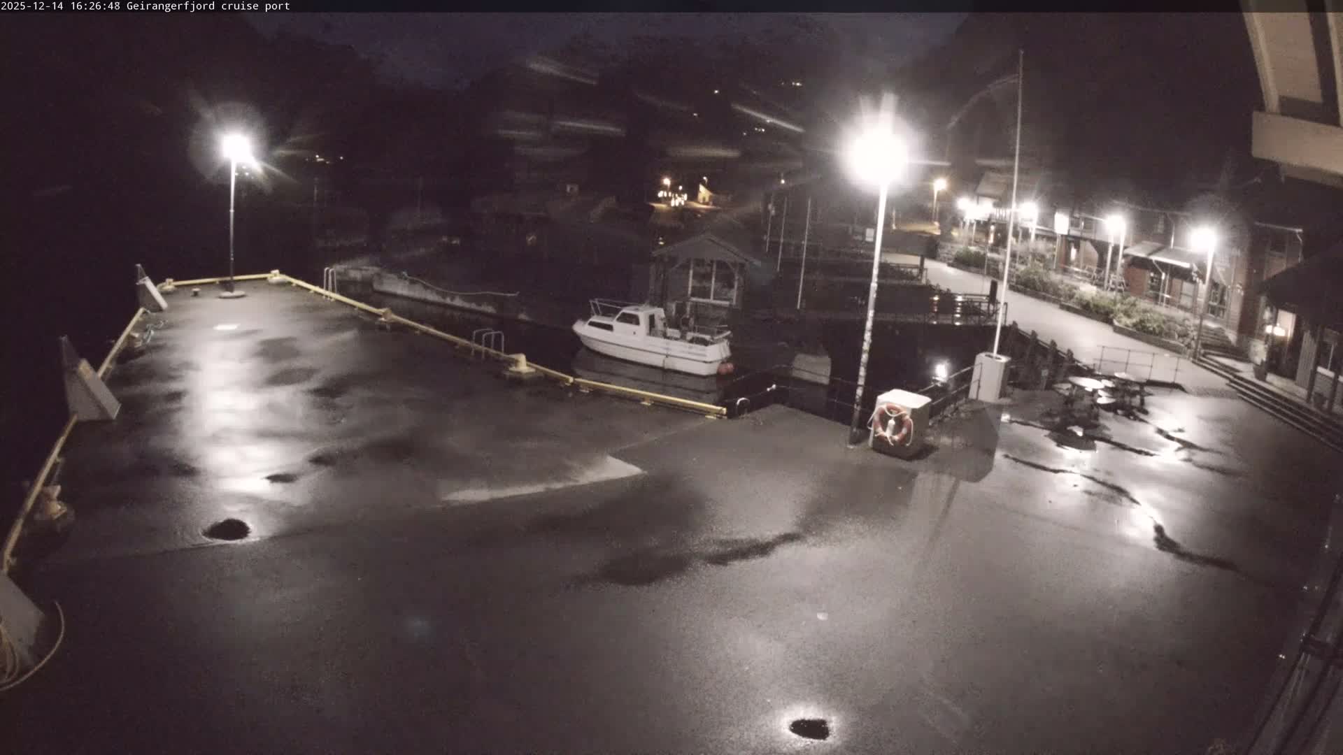 A serene winter landscape depicts a fjord port with dark, calm water reflecting snow-capped mountains, featuring a small boat docked at a pier adorned with piles of snow, and several waterside buildings under an overcast and cold sky.