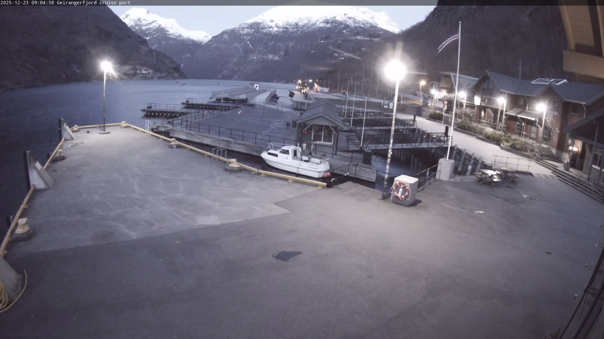 An early morning view of a quiet fjord port features a wide concrete pier with a white motorboat docked, traditional illuminated wooden buildings along the right side, and majestic snow-capped mountains rising from the water under an overcast sky.