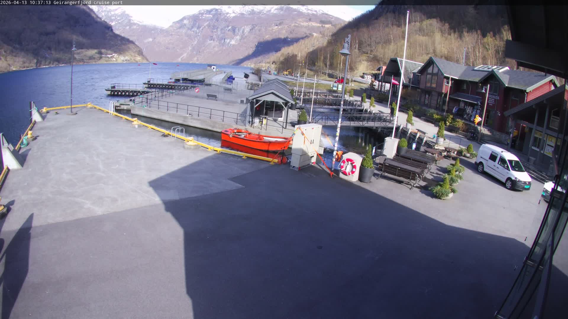 An early morning view of a quiet fjord port features a wide concrete pier with a white motorboat docked, traditional illuminated wooden buildings along the right side, and majestic snow-capped mountains rising from the water under an overcast sky.