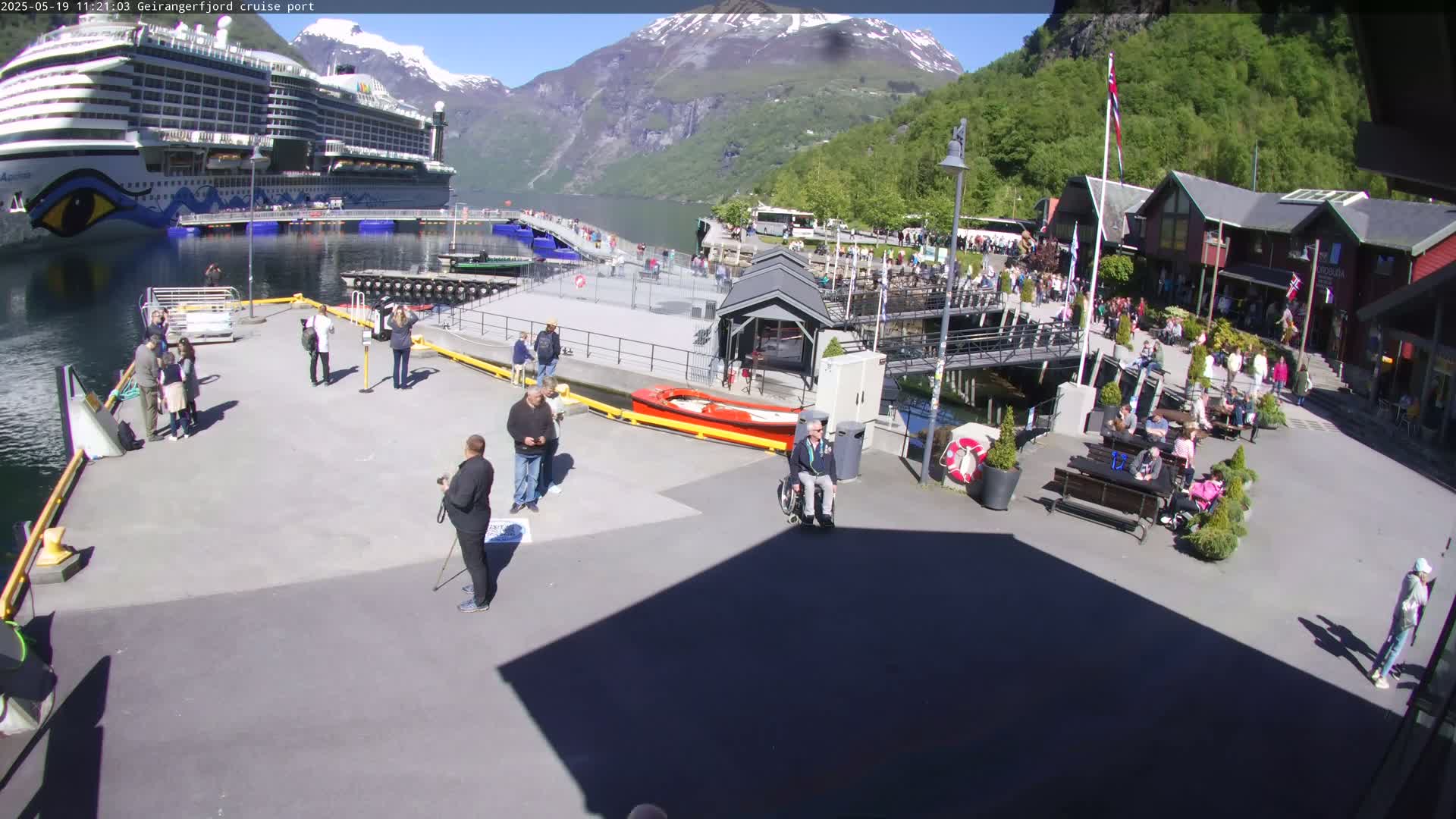 A large cruise ship is docked at a port in a fjord surrounded by mountains under a sunny sky, with many people walking around the port area.