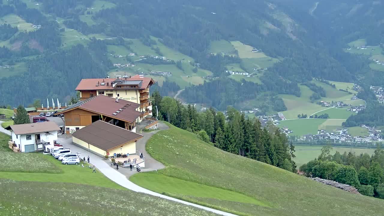 A mountaintop lodge with a parking lot and several smaller buildings overlooks a valley of green fields and dark evergreen forests on an overcast day.