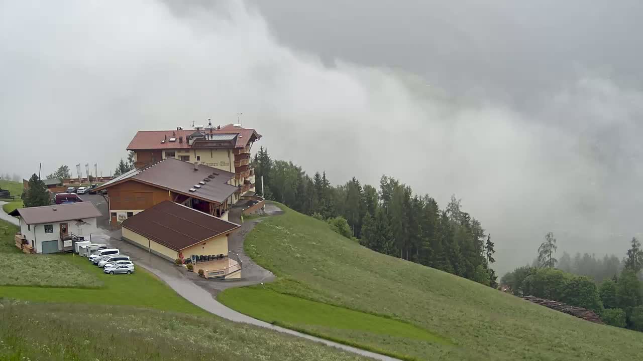 A multi-building complex sits on a grassy hillside overlooking a foggy forest under an overcast sky.