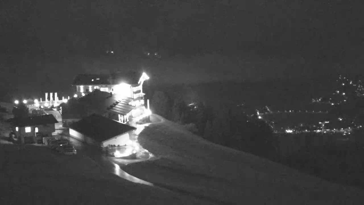 A nighttime, snow-covered hillside scene features a lit building complex, a few vehicles in a parking area, and a distant, faintly lit town.