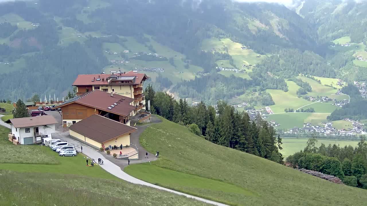 A large hotel complex sits atop a grassy hillside overlooking a valley of green fields and forests on a cloudy day.