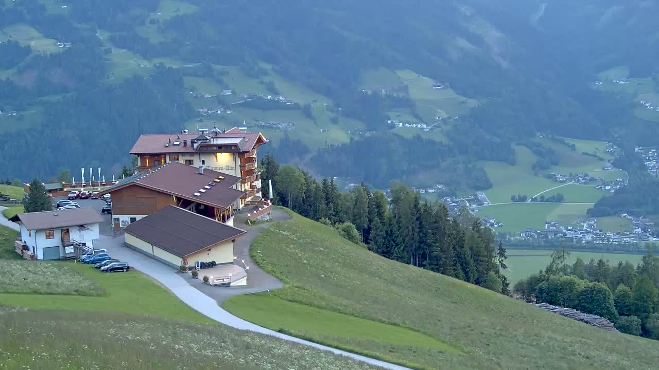 A mountaintop hotel complex with a parking lot overlooks a valley of green fields and a village under a hazy sky.