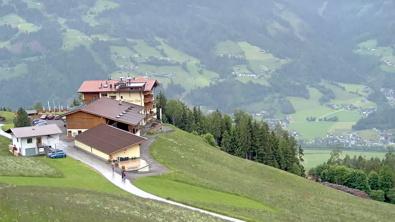 A mountaintop lodging complex with several buildings sits on a grassy hillside overlooking a valley of farmland and villages under an overcast sky.
