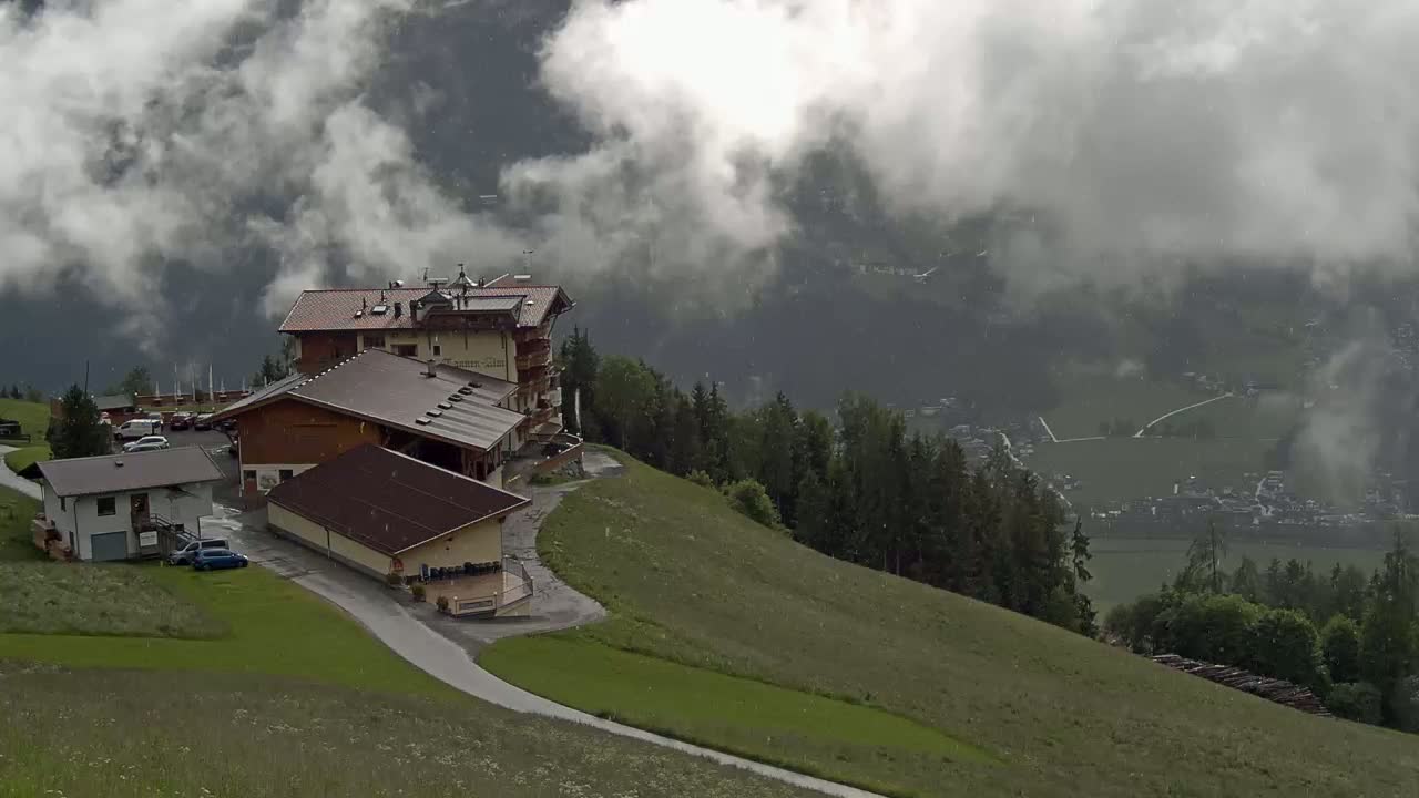 A large building complex sits atop a grassy hill overlooking a valley shrouded in mist and light rain.