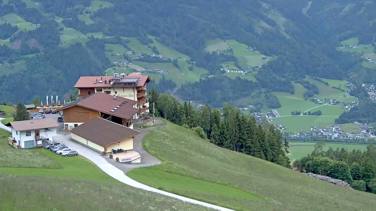 A brown multi-story building and several other smaller structures are situated on a grassy hillside overlooking a valley dotted with houses and farmland, under a mostly cloudy sky.