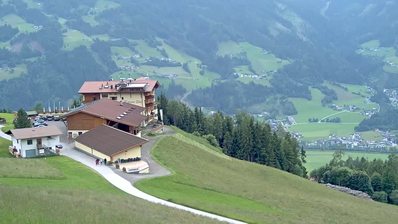 A complex of buildings sits atop a grassy hillside overlooking a valley of green fields and distant villages under a hazy sky.
