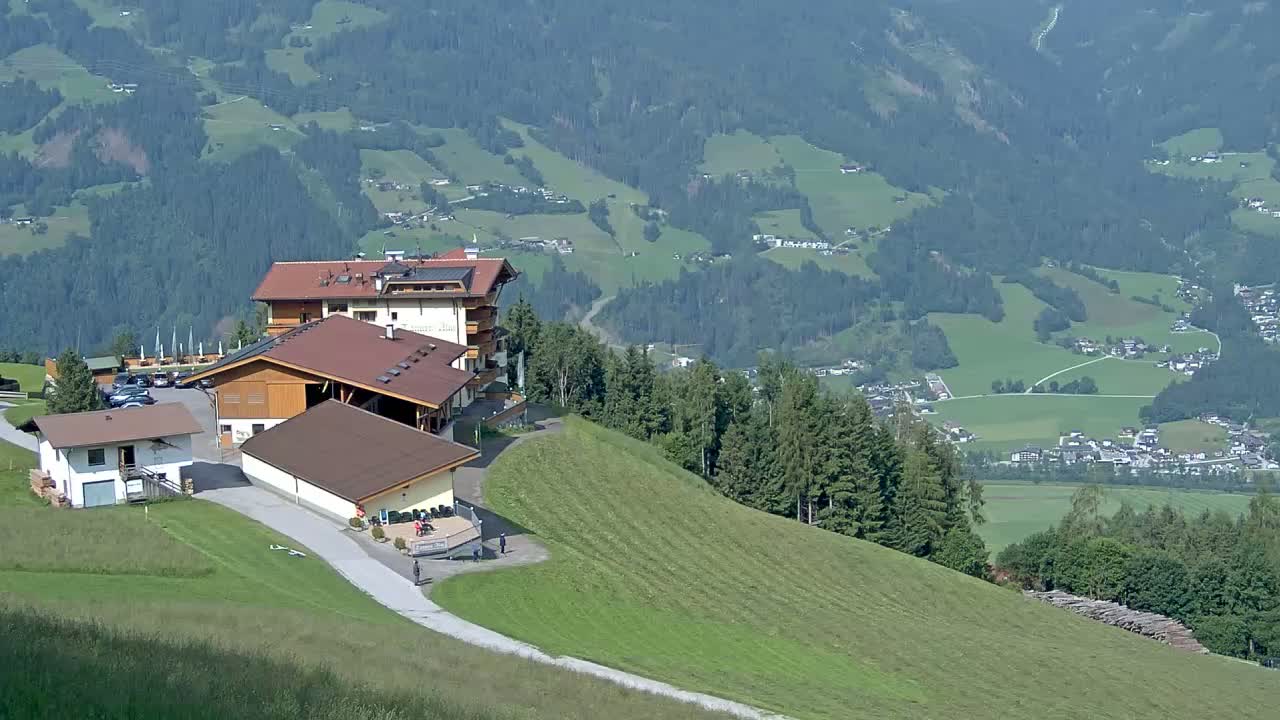 A mountaintop lodge with several buildings sits on a grassy hill overlooking a valley of green fields and distant villages under a clear sky.