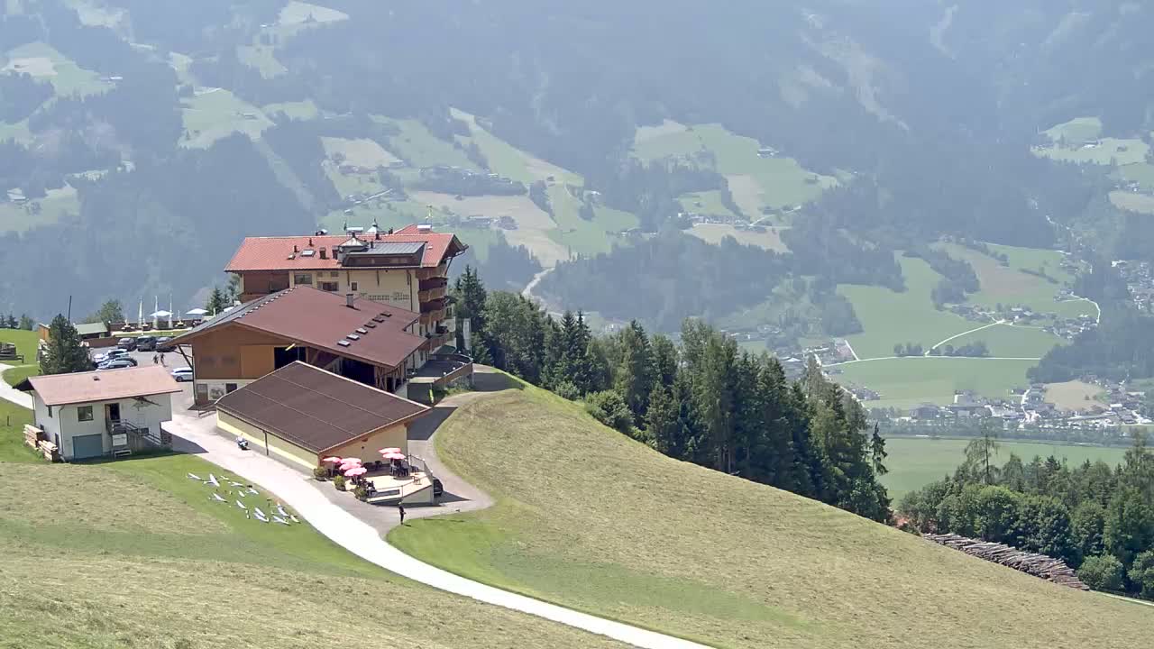 A mountaintop building complex overlooks a hazy valley of green fields and a distant village under a sunny sky.