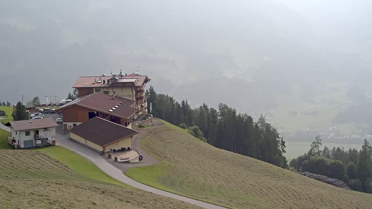 A hotel and several outbuildings sit on a grassy hillside overlooking a hazy valley and forest.