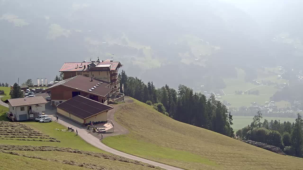 A mountaintop lodge and outbuildings sit on a grassy hillside overlooking a hazy valley with distant villages and forests.