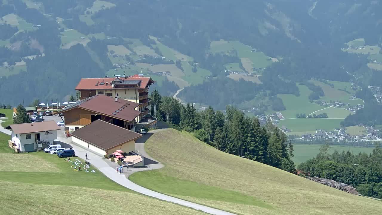 A mountaintop vista on a clear day shows several buildings, a paved road, and a grassy hillside overlooking a valley with rolling green fields and a distant village.