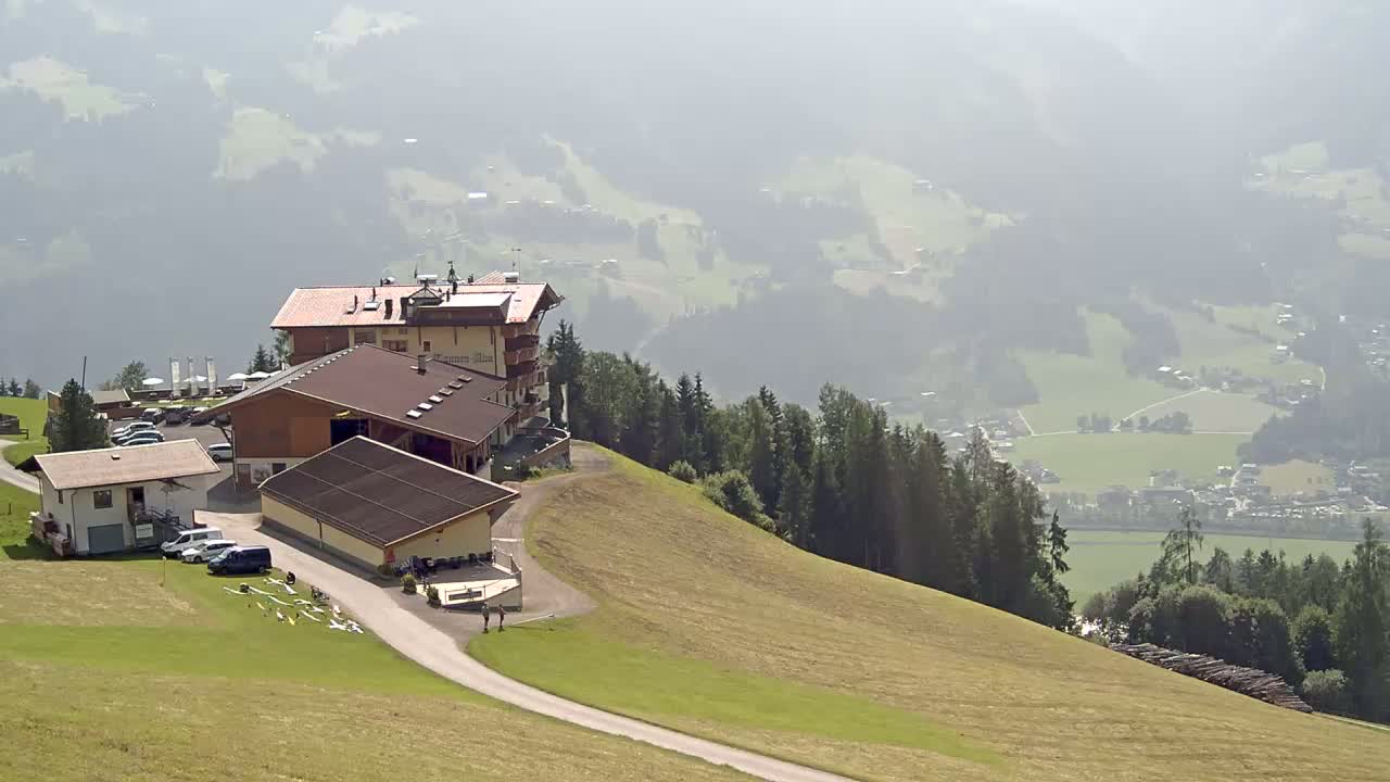 A mountaintop complex of buildings, including a hotel and outbuildings, overlooks a hazy valley with a village nestled amongst green fields and forests.